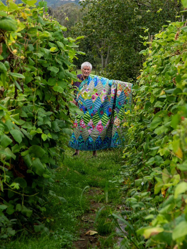 Kaffe Fassett pitelee ‘Flowers and Fences’ -tilkkupeittoa käsissään vihreässä puutarhassa, esitellen tilkkupeiton värikästä kukkakuviota ja maalaisaihetta.